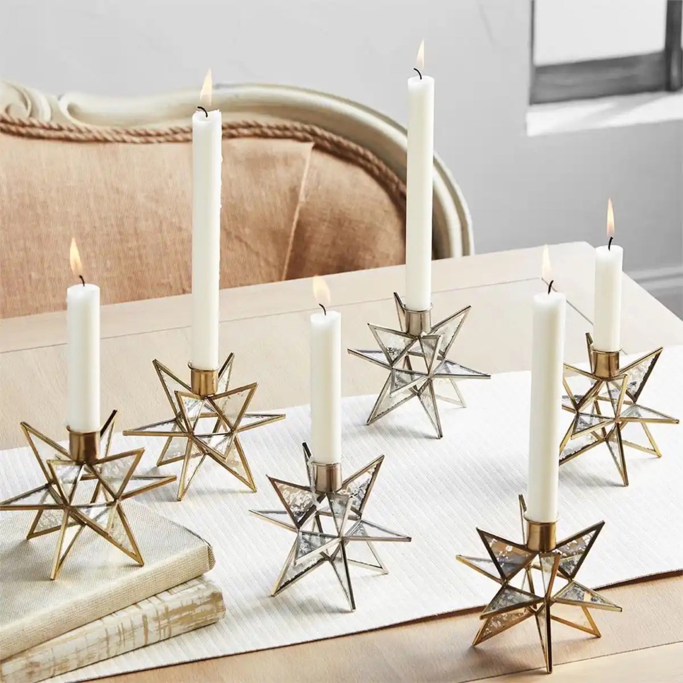 Set of three gold metal star candle holders with mercury glass panels holding white taper candles on white surface with books and basket
